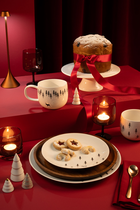 Une table dressée pour une fête avec un grand gâteau, entouré d'un ruban rouge et saupoudré de sucre glace, sur un présentoir. Il y a des biscuits sur des assiettes, des gobelets à thème, des bougies, des décorations de Noël rouges et blanches et un fond rouge foncé.
