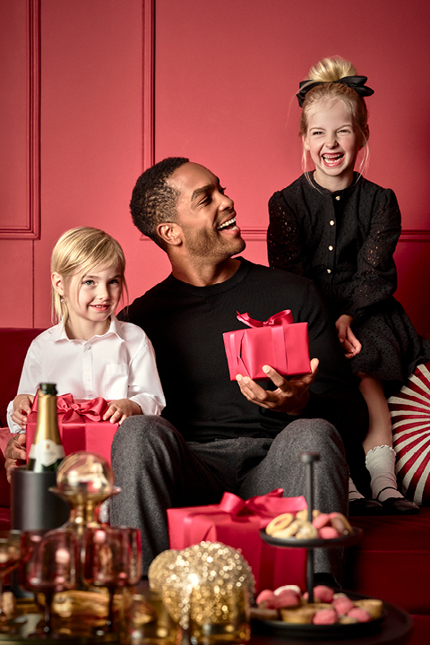 Un homme est assis sur un canapé rouge avec une boîte cadeau rouge et sourit à deux jeunes enfants. Un enfant, vêtu de blanc, tient un cadeau rouge similaire, tandis que l'autre, vêtu de noir, est assis sur le dossier du canapé. Au premier plan, il y a une table avec des friandises et une bouteille.