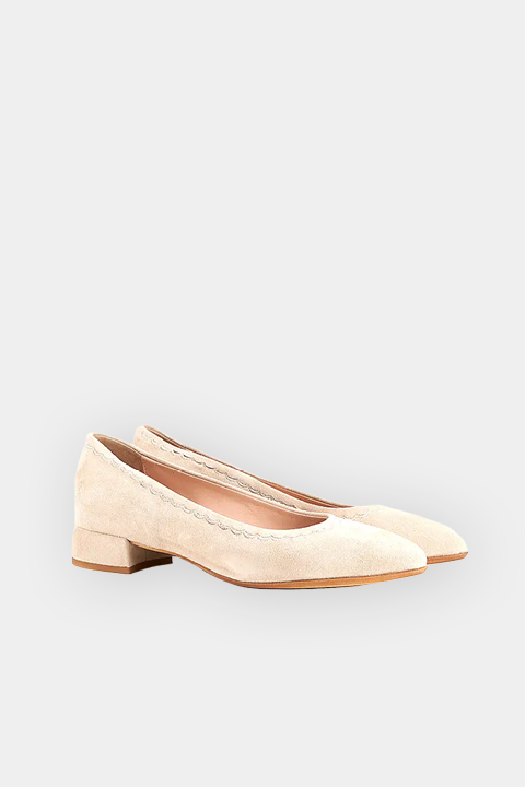 Une paire de chaussures basses en daim beige à bouts pointus, bords dentelés et semelles marron, présentées sur un fond gris clair.