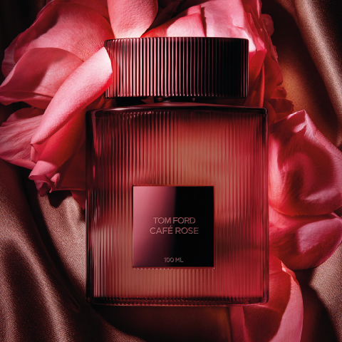 Une bouteille du parfum Tom Ford Café Rose est entourée de pétales de roses roses sur un tissu soyeux bronze. La bouteille est rectangulaire avec des rainures verticales et une étiquette sombre et carrée au centre, évoquant le luxe et l'élégance.