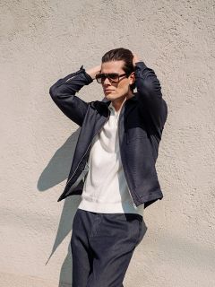 Un homme portant des lunettes de soleil, une veste bleu marine et un pantalon assorti se tient debout, les mains sur la tête, devant un mur clair et texturé.