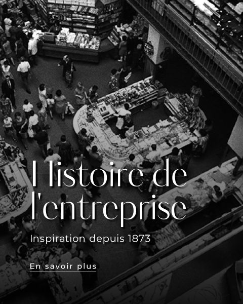 Photo en noir et blanc d'un marché couvert animé ou d'un grand magasin avec des personnes faisant leurs achats à différents comptoirs. Le texte superposé indique : "Histoire de l'entreprise Inspiration depuis 1873."