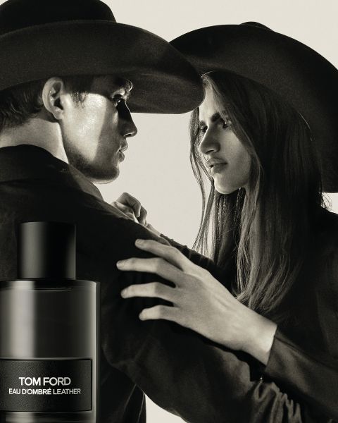 Un homme et une femme portant des chapeaux à larges bords se font face avec des expressions tendues. Au premier plan, on aperçoit un flacon d'Eau d'Ombré Leather de Tom Ford. L'image est en noir et blanc, ce qui lui confère une impression dramatique et classique.