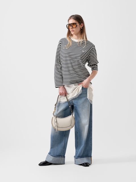 Femme portant un pull à rayures noires et blanches, un jean à jambes larges, des lunettes de soleil et un sac à main blanc devant un simple fond blanc.
