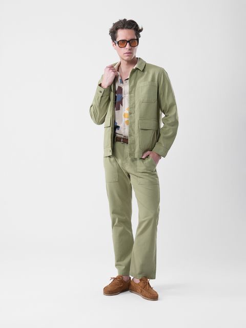 Un homme portant des lunettes de soleil, une veste et un pantalon vert clair, une chemise à motifs et des chaussures marron se tient devant un simple fond blanc.