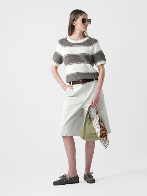Femme portant des lunettes de soleil, un pull rayé à manches courtes, un short blanc au genou et des chaussures basses, tenant un sac à main vert avec un foulard à la main.
