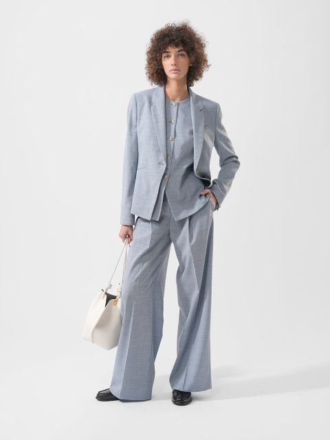 Une femme aux cheveux bouclés porte un tailleur gris clair ample, des chaussures noires et tient un sac à main blanc devant un fond blanc uni.