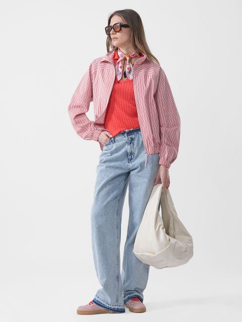 Une femme portant des lunettes de soleil porte une veste rayée rose, un pull rouge, un jean bleu et une écharpe, et tient un grand sac blanc devant un fond blanc uni.