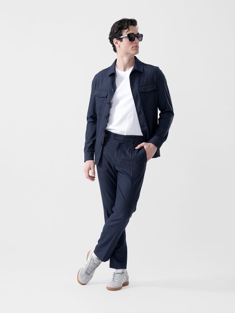 Un homme portant des lunettes de soleil noires, une veste bleu marine, un t-shirt blanc, un pantalon bleu marine et des baskets grises, se tient debout avec un pied croisé devant l'autre et regarde sur le côté.