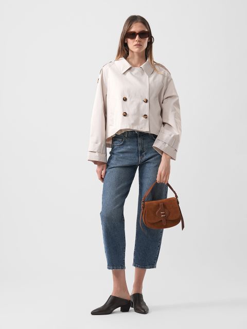 Une femme portant des lunettes de soleil porte une veste croisée beige, un jean bleu coupé, des chaussures noires et tient un sac à main marron devant un fond uni.