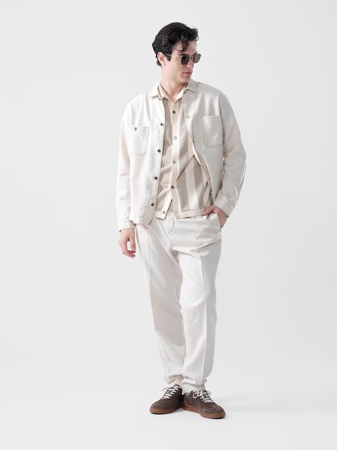 Un homme portant des lunettes de soleil, une veste beige clair, une chemise rayée et un pantalon assorti se tient debout, les mains dans les poches, devant un simple fond blanc.