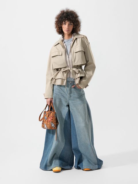 Une personne portant une veste beige, un jean bleu ample et des chaussures jaunes, tenant un sac à main à motifs marron, se tient devant un fond blanc.