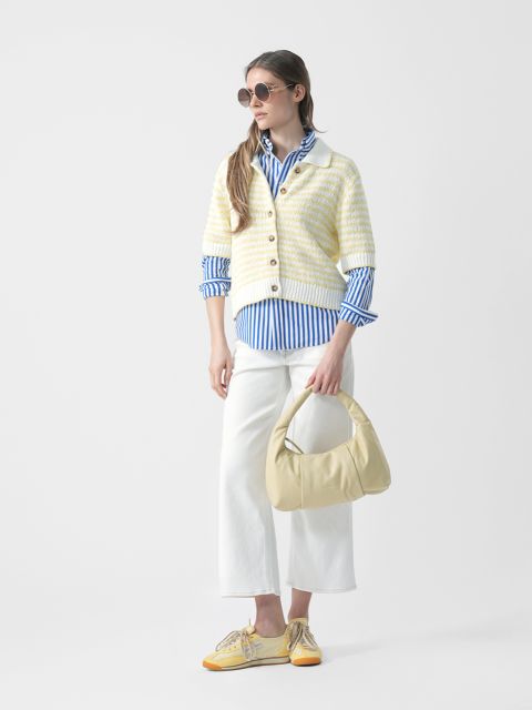 Femme avec des lunettes de soleil, une chemise bleue rayée, un cardigan rayé, un pantalon blanc et des chaussures jaunes tient un sac à main jaune clair et se tient devant un fond blanc.
