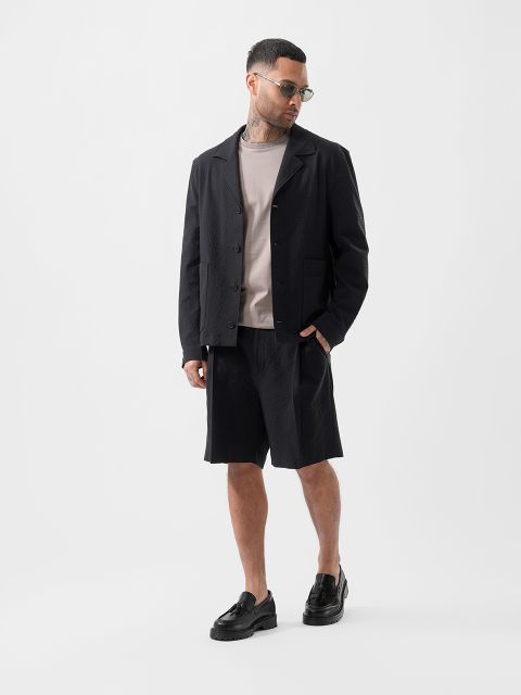 Un homme portant des lunettes de soleil, une veste noire texturée, un t-shirt beige, un short noir et des chaussons noirs se tient devant un simple fond blanc.