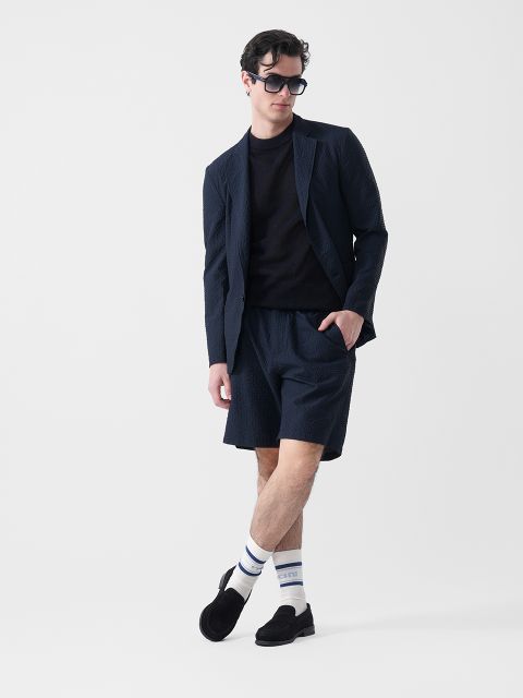 Un homme portant un blazer foncé, un short assorti, un t-shirt noir, des lunettes de soleil, des chaussettes blanches à rayures et des chaussons noirs se tient devant un fond blanc.