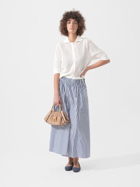 Femme portant un haut en tricot blanc à manches courtes, une jupe maxi rayée bleu et blanc, des ballerines bleu marine et un sac à main beige devant un fond uni.