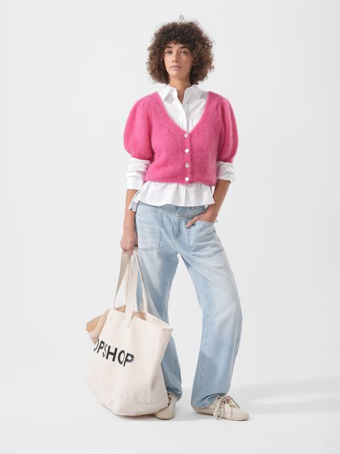 Une personne portant un cardigan rose, une chemise blanche, un jean bleu clair et des baskets tient un grand sac fourre-tout beige avec l'inscription "TOPSHOP".