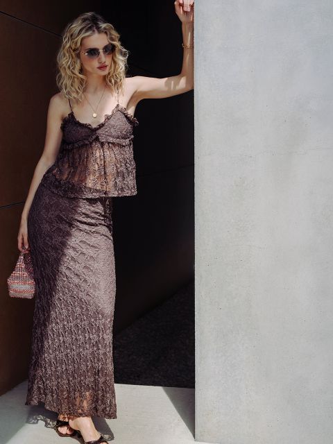 Une femme aux cheveux blonds bouclés, portant des lunettes de soleil et une robe en dentelle marron, s'appuie contre un mur en plein soleil, un petit sac à main à la main.