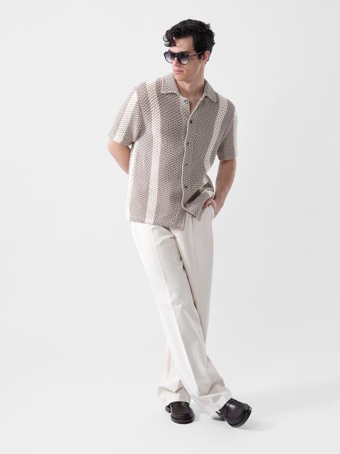 Un homme avec des lunettes de soleil, une chemise à manches courtes beige à motifs et un pantalon crème, se tient une main dans la poche devant un fond uni.