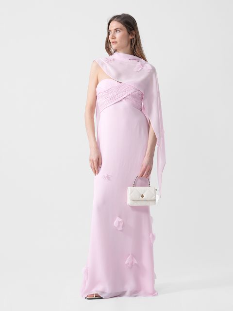 Une femme vêtue d'une robe bustier rose clair avec des détails floraux tient un petit sac à main blanc et porte un châle transparent assorti sur une épaule.