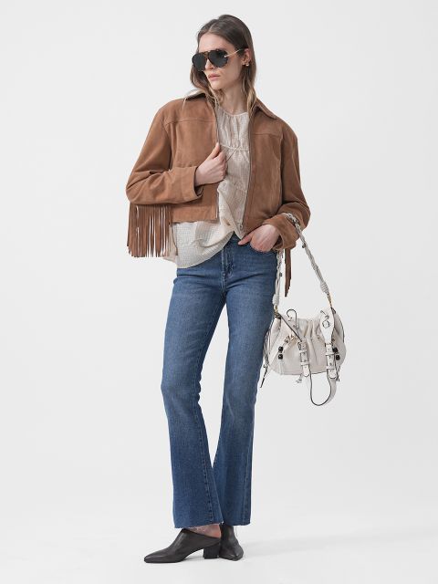 Une femme portant des lunettes de soleil, une veste marron à franges, un haut beige, un jean bleu, des chaussures noires et un sac à main blanc, pose devant un fond blanc.