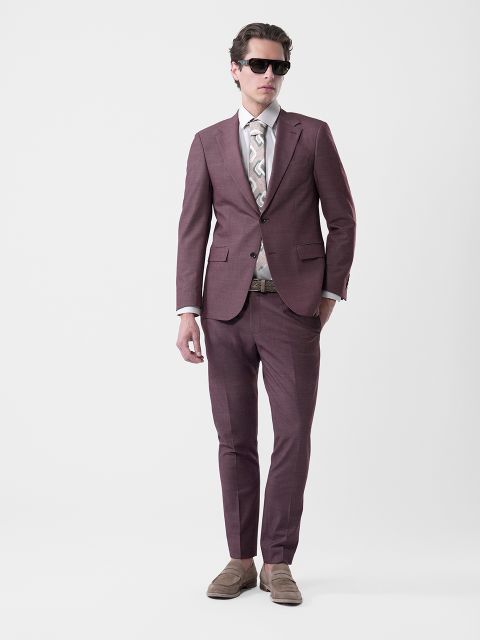 Homme en costume violet avec une cravate à motifs, une chemise blanche, des lunettes de soleil et des chaussures basses beiges devant un fond blanc uni.