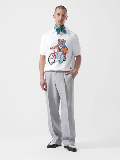 Un homme se tient devant un fond blanc, vêtu d'un pantalon gris clair, d'une chemise blanche avec un graphique d'ours, de chaussures bicolores et d'une écharpe bleu-vert nouée autour de son cou.