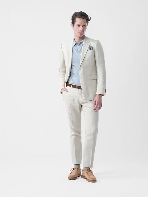 Homme en costume crème, chemise bleue, ceinture marron et chaussures marron, debout devant un fond blanc uni et regardant l'objectif.