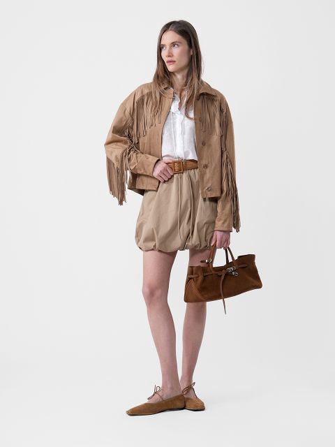 Femme en veste à franges marron clair, chemisier blanc, short marron clair avec ceinture, chaussures plates marron et sac à main marron devant un fond uni.
