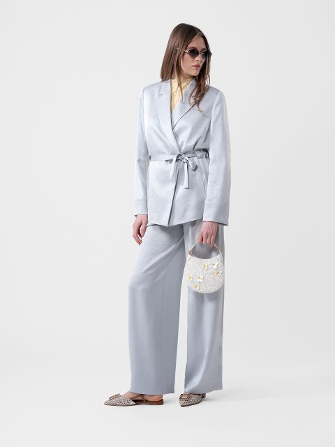 Femme vêtue d'un costume gris clair avec un blazer ceinturé, une chemise jaune, des lunettes de soleil, un petit sac à main blanc et des chaussures plates à motifs.