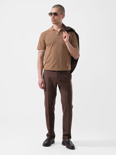 Un homme se tient devant un fond blanc, porte des lunettes de soleil, un polo marron, un pantalon marron foncé et tient une veste sombre sur l'épaule.