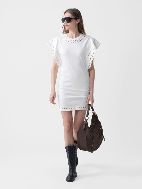 Femme en robe blanche à manches bouffantes, lunettes de soleil noires, bottes noires et grand sac à main marron, marchant devant un fond uni.