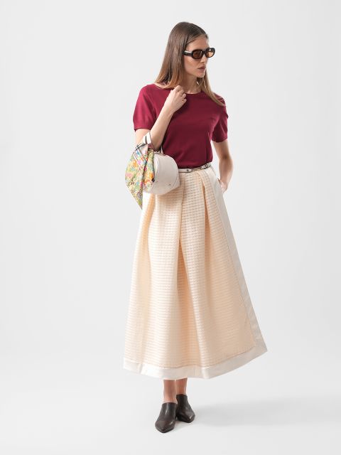 Femme portant un haut rouge, une longue jupe crème, des chaussures noires, des lunettes de soleil et tenant un sac à main à motifs, debout devant un fond uni.