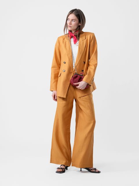 Femme en tailleur jaune moutarde, haut blanc, écharpe rouge, avec un petit sac rouge et des sandales noires, devant un simple fond blanc.