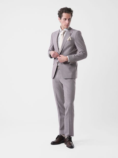 Un homme en costume gris clair, chemise crème et cravate, debout devant un fond blanc uni, boutonnant sa veste et regardant sur le côté.