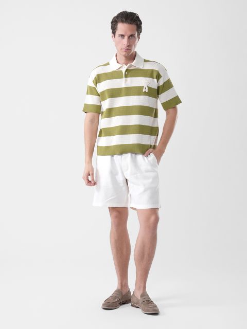 Homme en polo rayé vert et blanc, short blanc et chaussures basses marron devant un simple fond blanc.