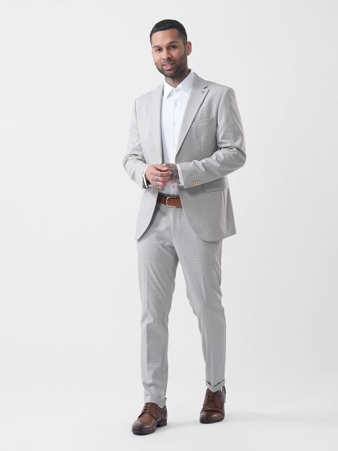 Homme en costume gris clair avec une chemise blanche et des chaussures marron, posant devant un simple fond blanc.