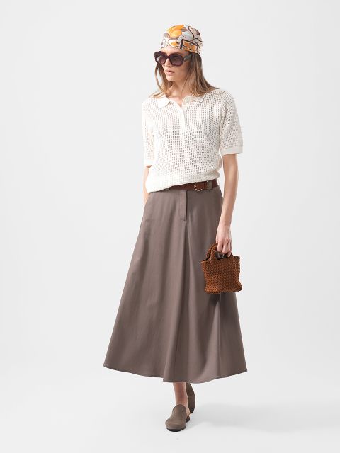 Frau trägt ein weißes, kurzärmeliges Strickoberteil, einen taupefarbenen Maxirock, ein Kopftuch, eine Sonnenbrille, einen braunen Gürtel, braune Schuhe und hält eine braune, gewebte Tasche.