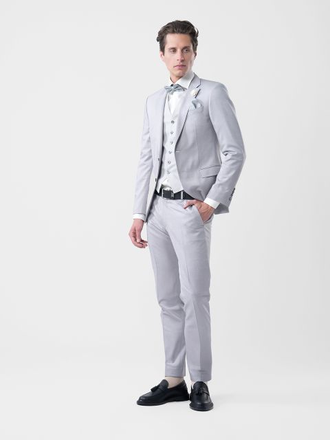 Un homme en costume gris clair avec une chemise blanche, un gilet à motifs et un nœud papillon, debout devant un simple fond blanc.