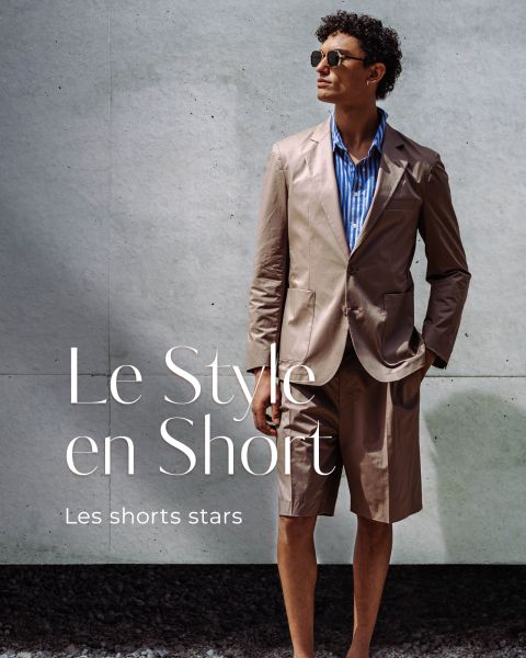 Un homme avec des lunettes de soleil porte un blazer marron clair et un short assorti par-dessus une chemise rayée bleue et se tient devant un mur en béton. Texte : "Le Style en Short. Les shorts stars.