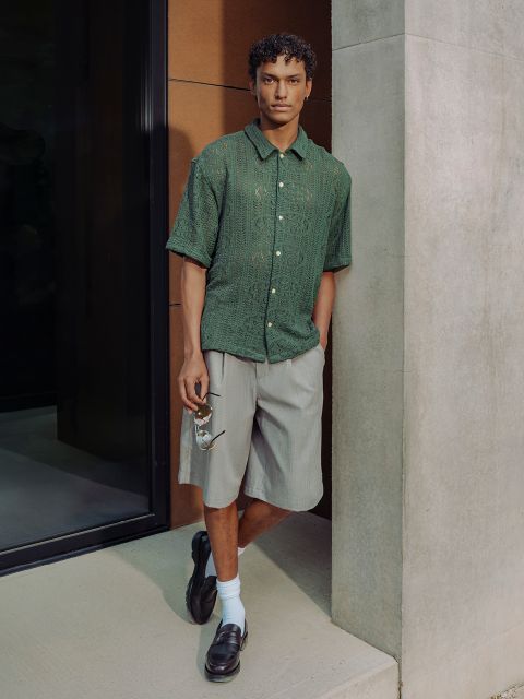 Un homme en chemise verte à manches courtes, short gris, chaussures basses noires et chaussettes blanches se tient contre un mur en béton et porte des lunettes de soleil.