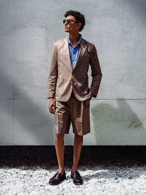 Un homme, portant des lunettes de soleil, un blazer beige clair avec un short assorti, une chemise rayée bleue et des mocassins, se tient devant un mur en béton sur du gravier.
