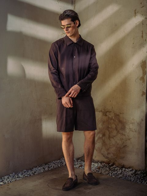 Un homme avec des lunettes de soleil, une chemise boutonnée marron foncé, un short assorti et des chaussures basses se tient devant un mur ensoleillé et texturé avec des accents de pierre.