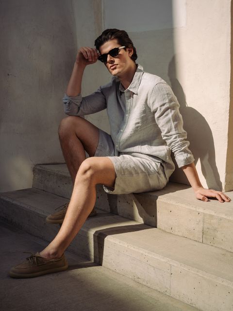 Un homme portant des lunettes de soleil, une chemise en lin claire et un short est assis sur un escalier en béton, au soleil, et appuie un bras sur son genou.