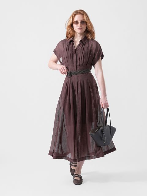 Une femme vêtue d'une robe marron transparente avec une ceinture, des lunettes de soleil et des sandales, portant un sac à main noir, marche avec assurance devant un arrière-plan blanc épuré.