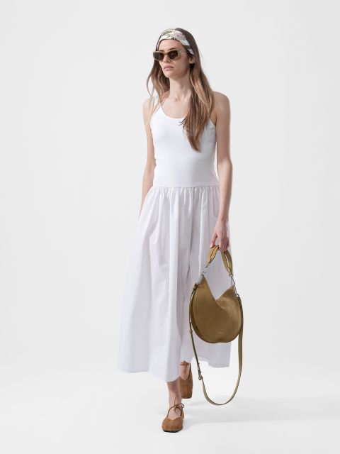 Femme portant une robe longue blanche sans manches, des lunettes de soleil, un foulard, des ballerines marron clair et un sac à main rond marron clair, devant un fond blanc uni.