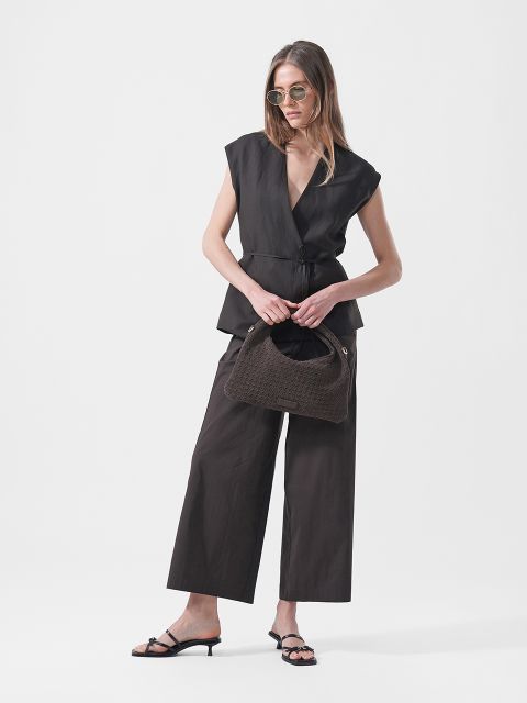 Une femme porte des lunettes de soleil, un haut cache-cœur noir sans manches, un pantalon noir à jambes larges et des sandales à talons hauts, et tient un sac à main texturé marron devant un fond blanc.