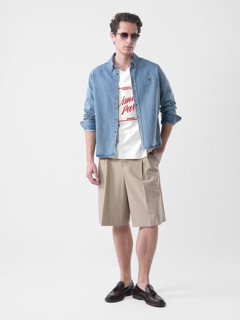 Un homme portant des lunettes de soleil, une chemise en jean sur un t-shirt graphique, un short beige au genou et des chaussures marron, debout devant un fond uni.