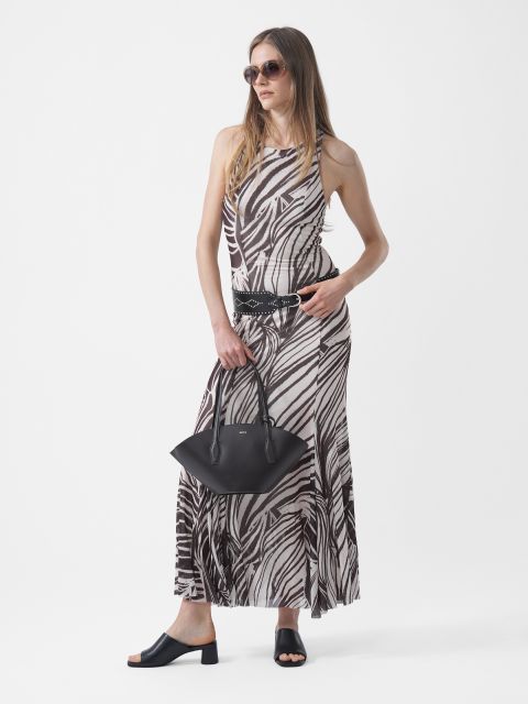 Femme portant des lunettes de soleil, vêtue d'une robe longue à motifs noir et blanc, d'une ceinture noire et de sandales noires, et tenant un sac à main noir devant un fond blanc.