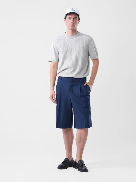 Un homme portant une casquette blanche, un pull à manches courtes gris clair, un short bleu marine arrivant aux genoux et des mocassins noirs se tient devant un fond blanc uni.